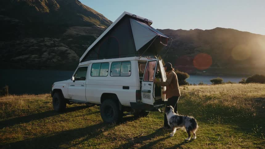 A man and his loyal dog stand by their pop-top camper van, overlooking a stunning lake and mountains at sunset