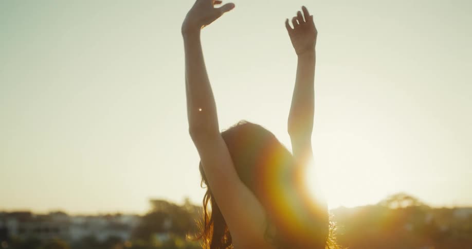 A young woman with long hair slowly dances with her arms raised against the golden setting sun, creating a beautiful silhouette and lens flare