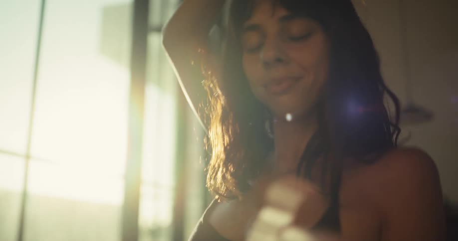A beautiful woman with eyes closed dances sensually in her sunlit apartment, moving gracefully in slow motion