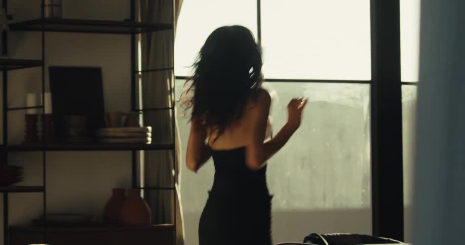 A joyful woman with dark hair dances freely in slow motion in her modern, sun-drenched living room