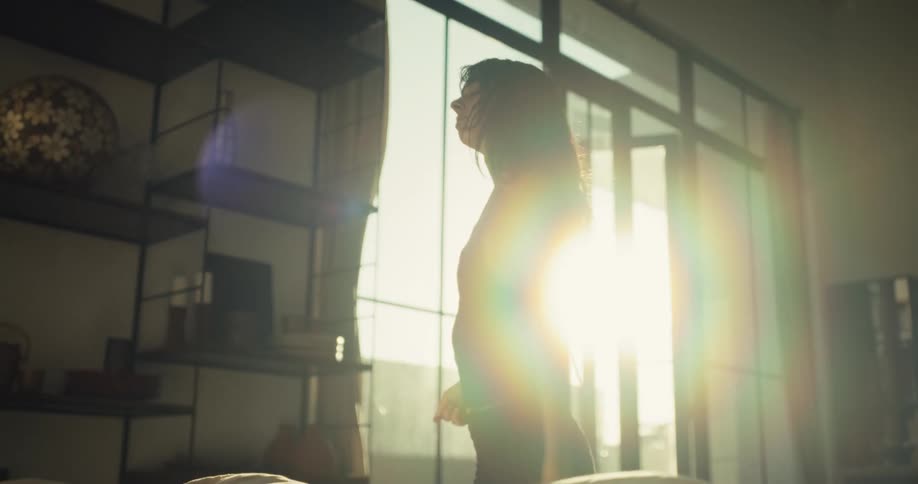 A young woman in her pajamas stretches and dances slowly as the morning sun streams through her bedroom window