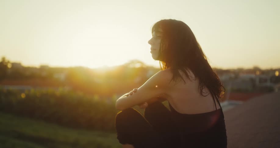 Rack focus shot of a thoughtful woman sitting on a rooftop enjoying the peaceful golden hour sunset over a tropical town