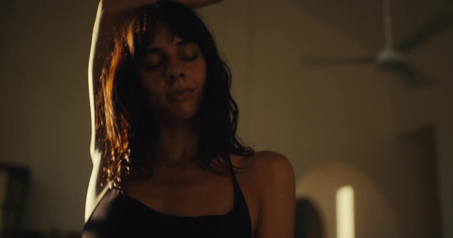 A young woman in a black top moves her body in a slow, sensual dance in a dimly lit room