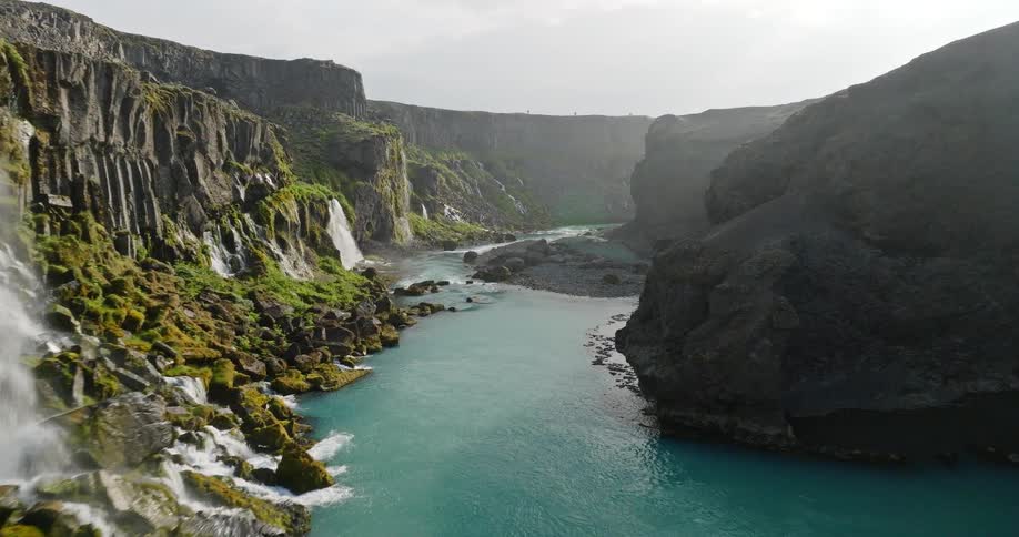 A breathtaking aerial flight through a rugged Icelandic canyon reveals a vibrant turquoise river and numerous waterfalls cascading down mossy cliffs