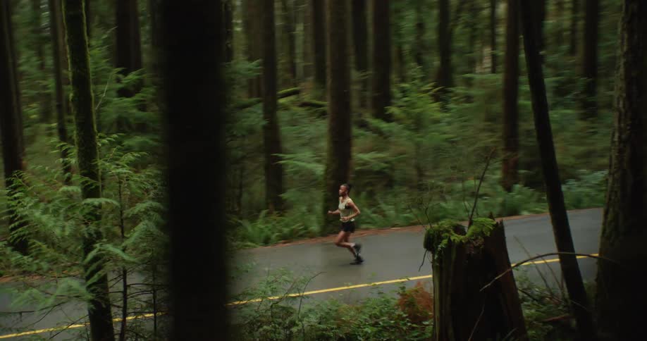 A determined female athlete runs along a winding paved road through a dense and lush green forest
