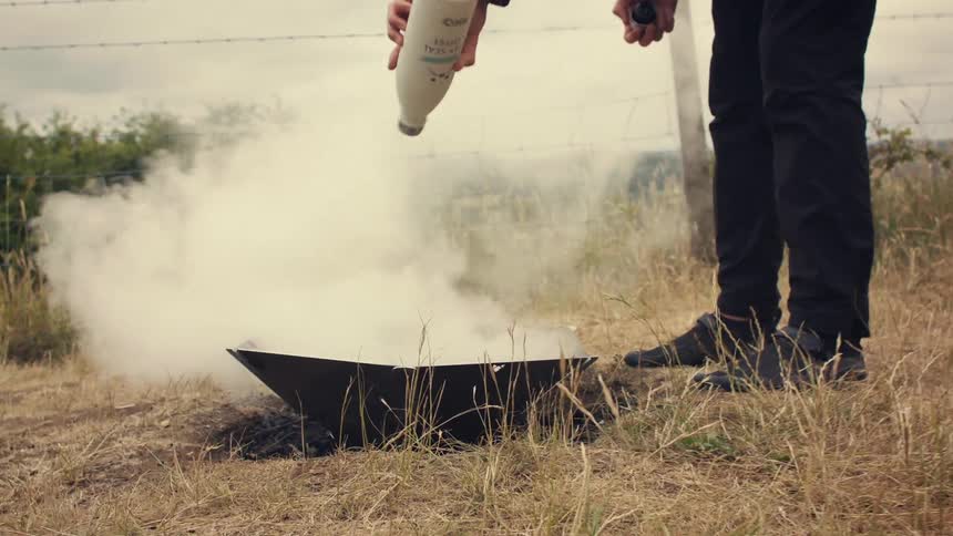A person pours liquid from a bottle into a smoking fire pit in a grassy, rural field