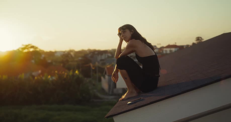 A pensive young woman in a black dress squats barefoot on a rooftop, watching the golden sunset over a tropical town