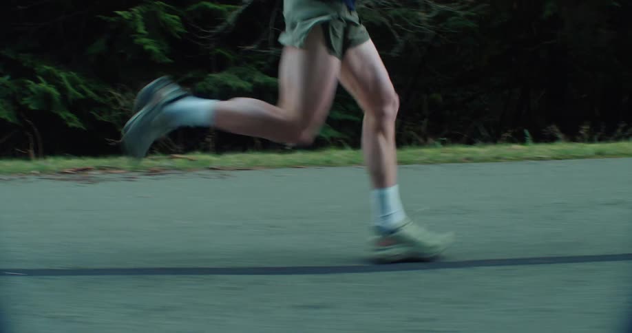 An athletic man with strong legs runs along a paved road in a park during an early morning workout