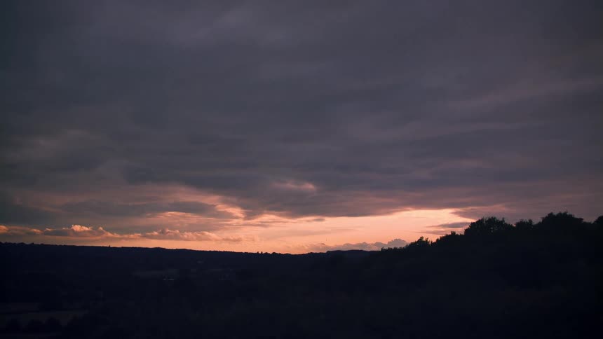 A dramatic sunset paints the horizon with orange light beneath a sky of dark, moody clouds, slowly panning over a silhouetted treeline