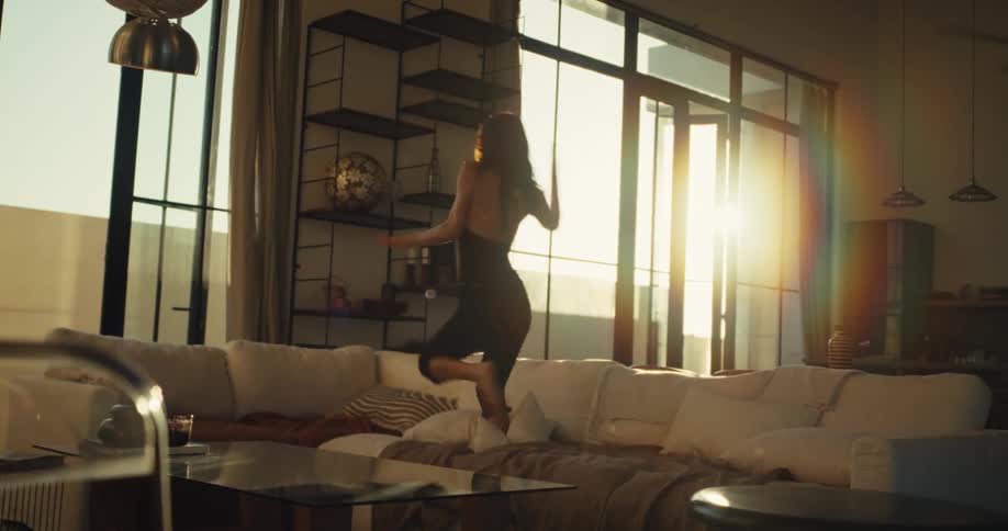 A carefree woman joyfully dances and jumps on her couch in a sun-drenched modern apartment, captured in slow motion