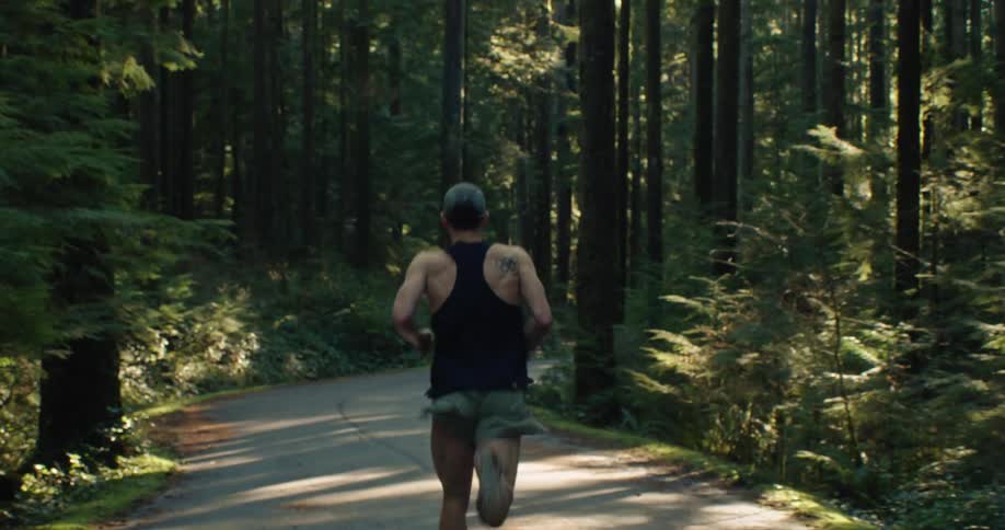 A fit man in a tank top and shorts jogs down a winding road through a sunlit evergreen forest