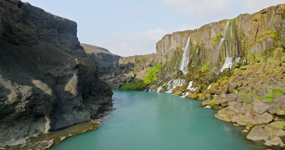 An aerial view follows two hikers to the edge of a breathtaking Icelandic canyon, soaring over to reveal a hidden oasis with a turquoise river and multiple waterfalls
