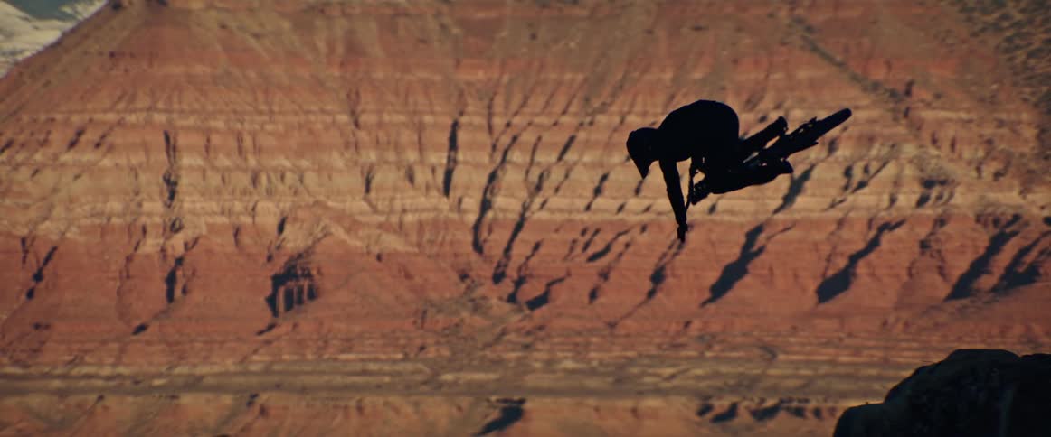 A silhouette of a mountain biker performs a tabletop trick in slow motion against a massive red rock canyon wall
