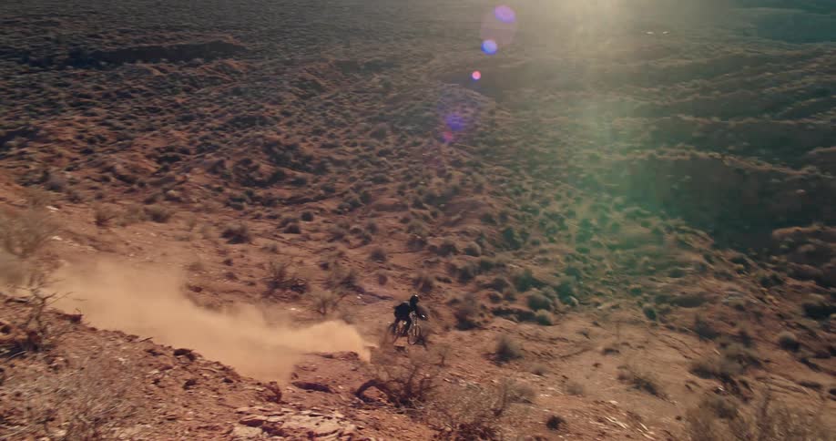 A mountain biker descends a steep, dusty red rock cliffside into a vast desert canyon