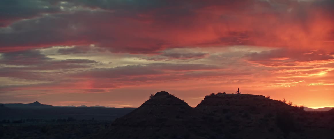 A silhouetted mountain biker rests on a desert butte, taking in the epic view of a fiery and dramatic sunset