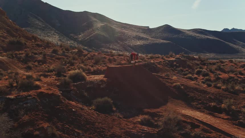 A mountain biker soars through the air off a dirt jump while performing a trick against a stunning desert mountain backdrop
