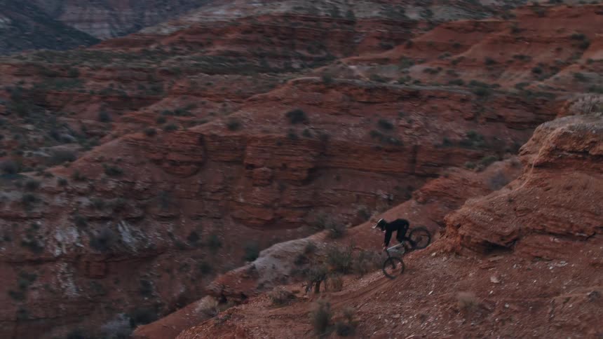 Aerial shot of a mountain biker carefully descending a steep and rugged red rock trail in a vast desert canyon