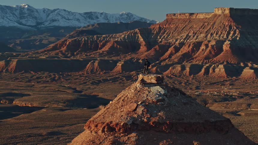 A lone mountain biker stands atop a desert butte, enjoying the epic view of red rock canyons and distant snowy mountains at sunrise