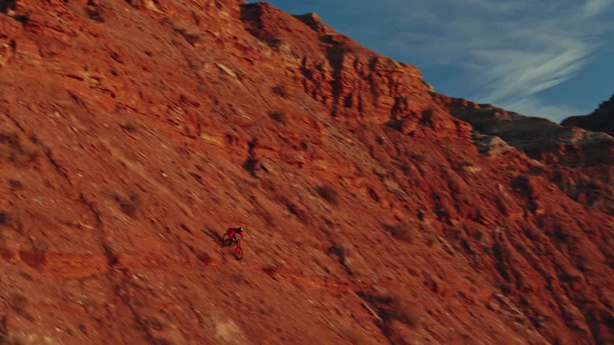 In a breathtaking aerial shot, a mountain bike athlete navigates down a steep, sunlit red rock mountain face at sunset
