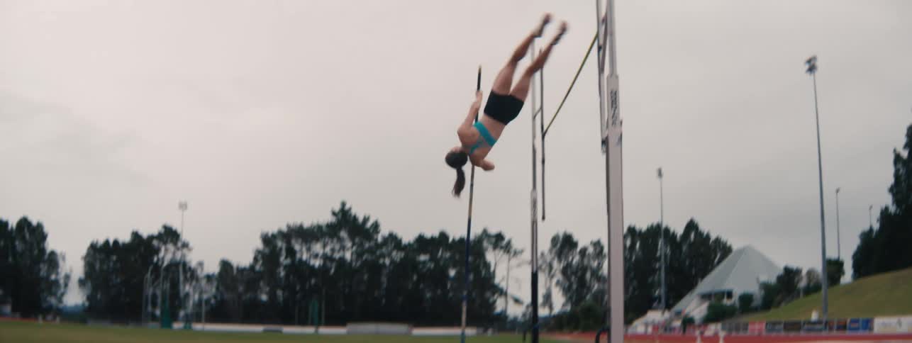 A female athlete gracefully pole vaults over the bar in slow motion at a track and field stadium