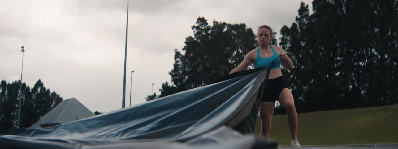 A determined female athlete pulls a heavy tarp across a sports field in slow motion