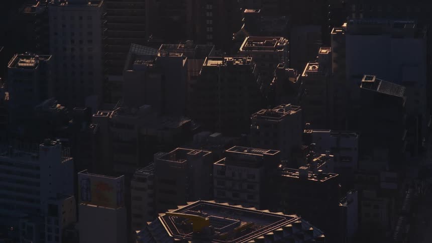Golden sunrise light slowly moves across the tops of buildings in a dense, shadowy urban cityscape