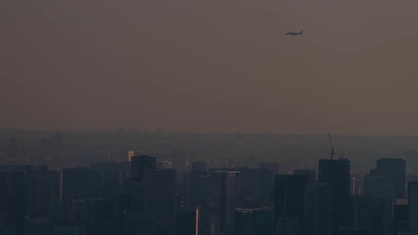 A passenger airplane flies over a vast, hazy city skyline at dusk