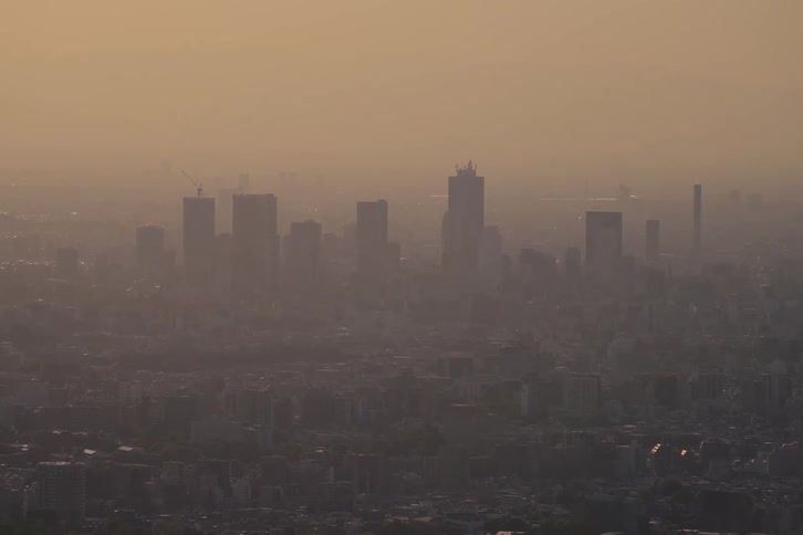 A static aerial view shows a polluted cityscape with skyscrapers rising through the thick orange haze at dusk