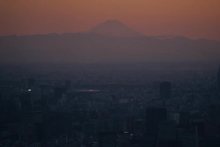The silhouette of Mount Fuji looms over the vast Tokyo cityscape during a hazy, colorful sunset