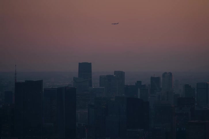 A commercial airplane flies high above a silhouetted city skyline at dusk