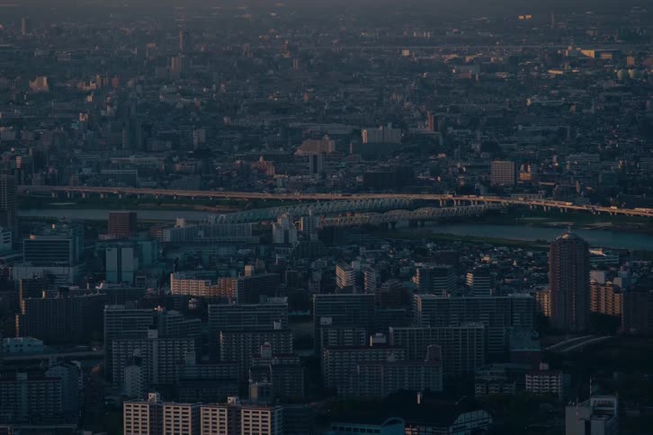 A serene timelapse captures the sprawling urban landscape of a major city at dusk, with traffic flowing over a bridge spanning a wide river