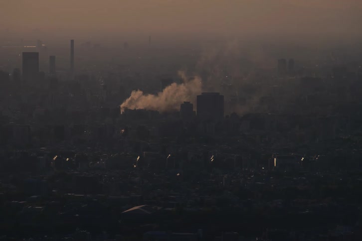 A plume of smoke rises from a fire in a vast, polluted city skyline at dusk