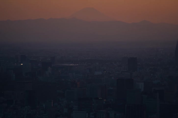 The majestic silhouette of Mount Fuji looms over the sprawling, hazy city of Tokyo during a warm sunset