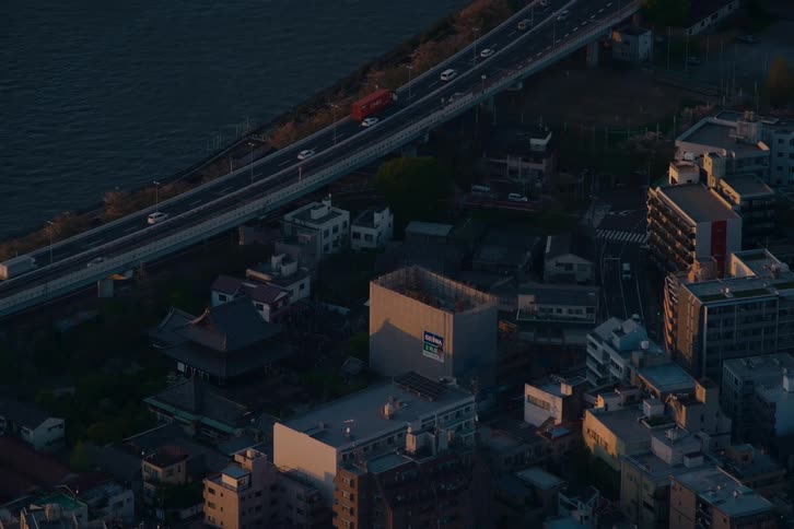 Aerial view of a coastal Japanese city at dusk, with traffic moving along an elevated highway next to a traditional temple and modern buildings