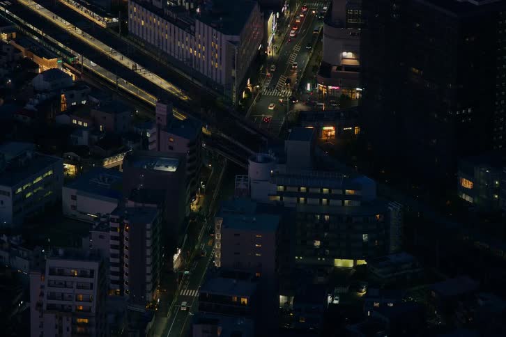An aerial view captures the bustling city of Tokyo at night, with trains arriving at the station and cars moving through the streets