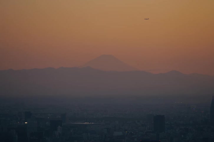 A serene view of Mount Fuji silhouetted against a vibrant orange sunset sky, with the sprawling cityscape of Tokyo below and an airplane flying overhead