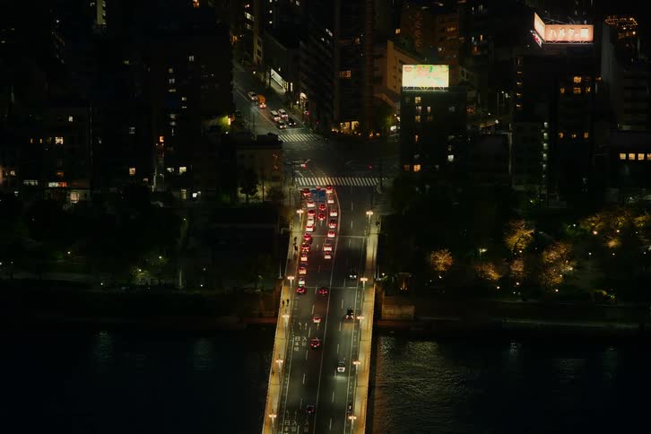A high-angle view shows evening traffic crossing a brightly lit bridge over a dark river in a bustling city at night