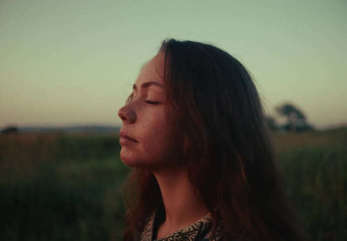 A thoughtful young woman with long hair and freckles takes a quiet moment to reflect in a field at sunset