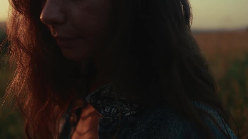 A close-up shot of a pensive woman with long hair standing in a field during the warm glow of golden hour