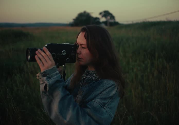 Cinematic shot of a young woman raising a vintage film camera to her eye in a grassy field at dusk