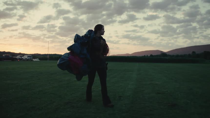 A silhouetted skydiver carries a bundled parachute while walking across a grassy field at sunset