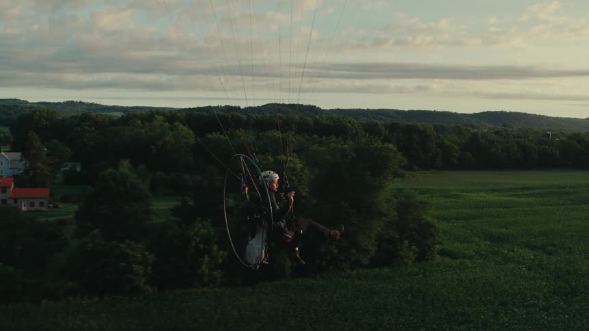 Slow motion aerial of a powered paraglider flying low over a lush green field in the countryside at sunset