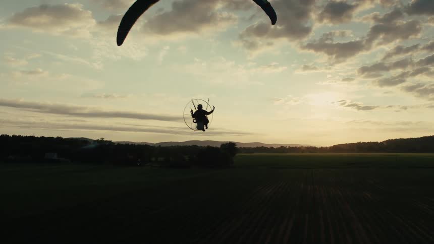 A stunning slow-motion aerial shot captures the silhouette of a powered paraglider flying through the sky at sunset