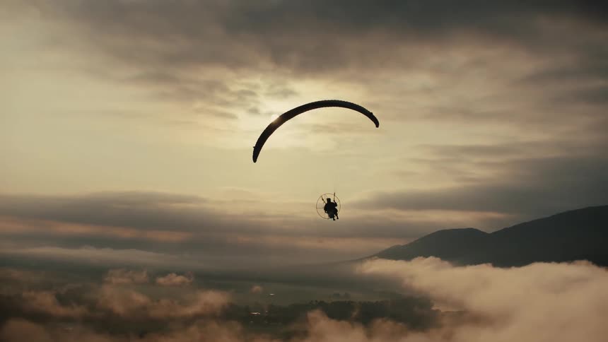 A silhouetted paraglider floats peacefully over a sea of clouds with mountains in the distance as the sun rises