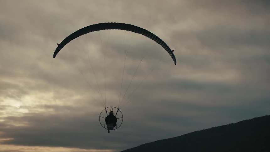 A silhouetted paramotor hovers peacefully in place against a cloudy sky at dusk