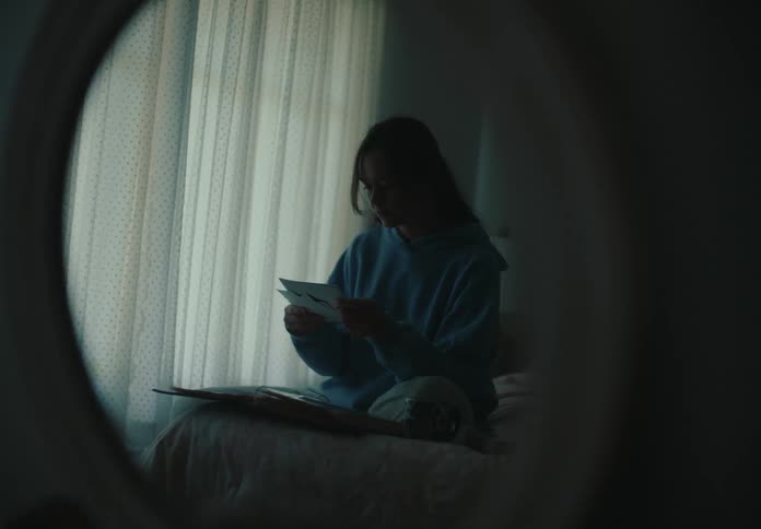 Reflection in a mirror shows a young woman sitting on her bed in a dimly lit room, reminiscing over old photographs