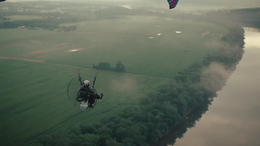 An aerial shot follows a powered paraglider as it soars through the morning mist above a wide river and verdant countryside