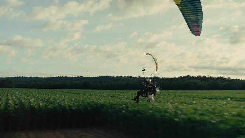 A slow-motion aerial tracking shot follows a powered paraglider as it soars low over a lush green field