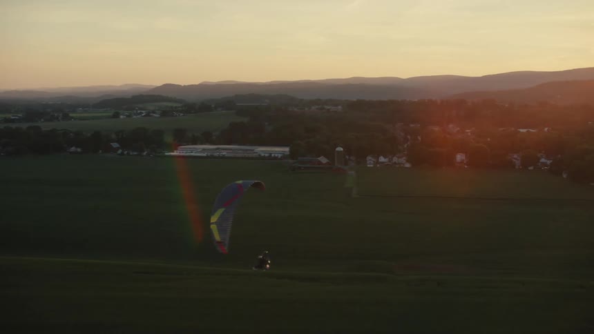 An aerial view of a powered paraglider soaring over lush green fields in the countryside during a beautiful sunset