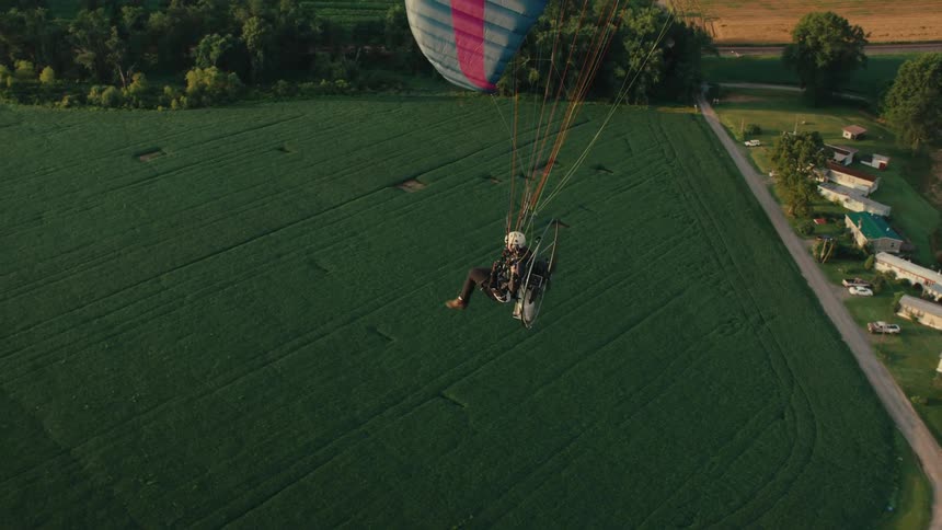 An aerial view captures a powered paraglider flying serenely over lush green fields and a rural town